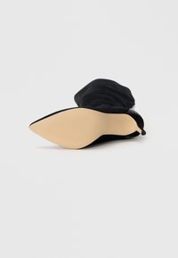 Bottine en suède noir avec un bout pointu et une semelle beige lisse. Présente un design de revers plié et un détail de talon texturé.