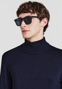 Schwarze Sonnenbrille mit quadratischen Rahmen, getragen über einem marineblauen Rollkragenpullover. Der Stoff wirkt glatt, mit einem figurnahen Design und geripptem Halsausschnitt.