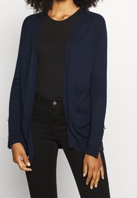 Námořnický pletený kardigan s otevřeným předním dílem, dlouhými rukávy a knoflíkovými detaily na manžetách. Kombinováno s černým přiléhavým topem a tmavými džínami.