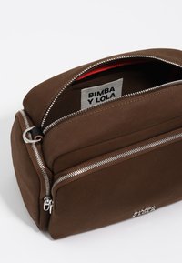 Rudas audinys krepšys su glotnia struktūra, turi sidabrinius užtrauktukus, raudoną vidinę pamušalą ir logotipo etiketę viduje. Stačiakampio formos su keliais skyreliais.