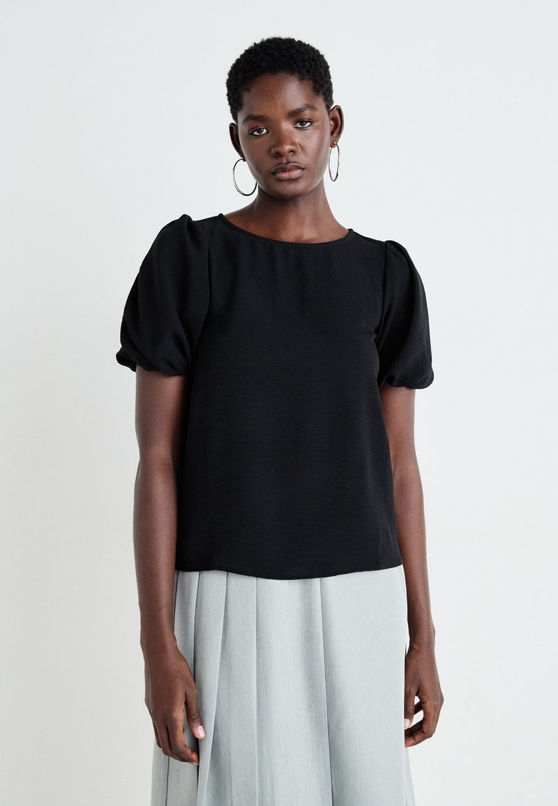 Blusa nera a maniche corte con scollatura rotonda e maniche a sbuffo gatherate; tessuto morbido, abbinata a una gonna plissettata grigia chiara.