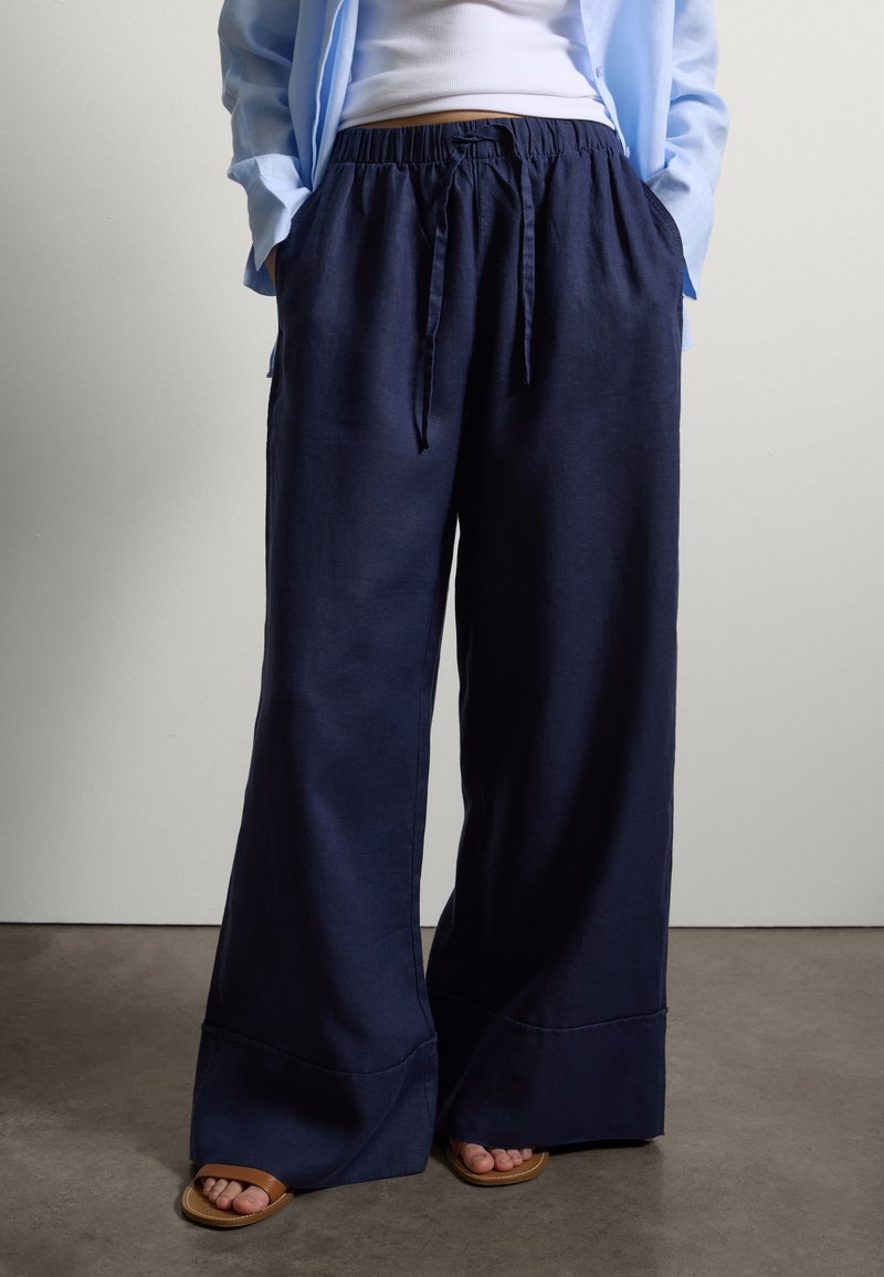 Person wearing navy wide-leg pants with drawstring waist, white tank top, light blue overshirt, and brown open-toe sandals standing on a gray floor.