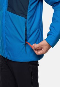 Blauwe buitenjas met een contrasterend zwart paneel, voorzien van een ritszak en een feloranje trekstang, gemaakt van lichtgewicht stof.