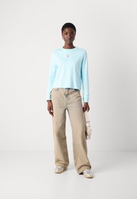 Helles blaues Langarmoberteil mit kleinem rotem Logo, kombiniert mit beigen Weitenjeans und silbernen Sneakers. Minimalistisches Design, lässige Passform.