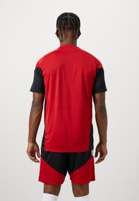 Κόκκινο και μαύρο σετ αθλητικής φανέλας και σορτς. Φανέλα κοντομάνικη με κόκκινο επάνω μέρος και μαύρα μανίκια, διαθέτει στρογγυλή λαιμόκοψη.