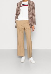 Pantaloni beige a gamba larga abbinati a un maglione bianco con strisce orizzontali rosse e blu navy, indossato sotto un blazer a quadri marrone. Sneakers bianche.