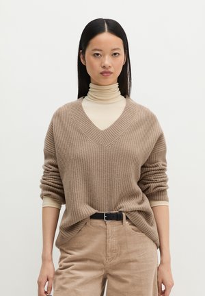 Jonge vrouw met steil zwart haar die een beige coltrui draagt onder een loszittende bruine trui met V-hals en bijpassende corduroy broek met zwarte riem.