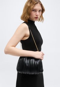 Γυναίκα με κοντά ξανθά μαλλιά που φοράει μαύρο αμάνικο φόρεμα, κρατώντας μια πλισέ μαύρη δερμάτινη τσάντα με χρυσή αλυσίδα.