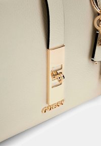 Sac à main en simili cuir beige avec fermoir en métal doré, logo gravé et accent de matériel décoratif. Surface texturée avec finition lisse.