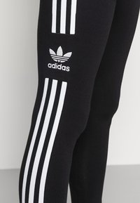 Leggings atletici neri caratterizzati da accenti bianchi a tre strisce che scendono lungo le gambe e un logo Adidas ricamato sulla coscia superiore.