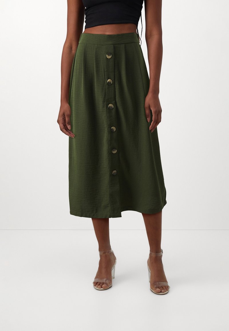 Femme portant une jupe olive longue jusqu’au genou avec de gros boutons devant et des sandales à talons hauts à brides transparentes.
