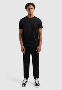 Zwart t-shirt met korte mouwen met een klein logo, gecombineerd met zwarte joggingsbroek en zwart-witte sneakers, weergegeven tegen een neutrale achtergrond.