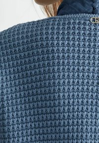 Vaaleansininen virkattu paita, jossa on teksturoitu ruudukkomainen kuvio. Kauluksessa nappikoriste ja alla sileää kangasta.