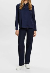 Mörkblå skjorta med krage, matchad med mörkblå utsvängda jeans och vita sneakers, stående mot en neutral bakgrund.