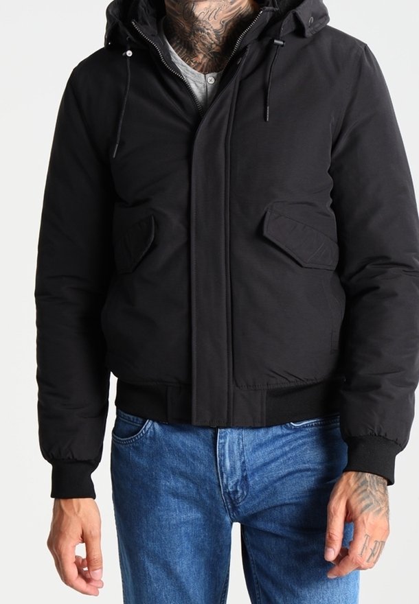 Homme portant une veste noire à capuche avec poches à rabat, poignets côtelés et jean bleu, montrant son cou et ses mains tatouées.