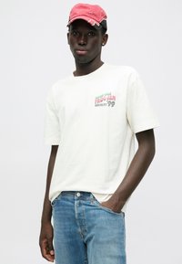 Λευκό βαμβακερό t-shirt με πολύχρωμο κείμενο σε πράσινο, ροζ και μαύρο χρώμα. Συνδυασμένο με ανοιχτό μπλε τζιν παντελόνι. Κόκκινο καπέλο του μπέιζμπολ στο κεφάλι.