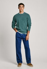 Zelena rebrasta pulover s krožnim ovratnikom, v kombinaciji z ohlapnimi denim hlačami in rjavimi čevlji, stoji pred enobarvno ozadjem.