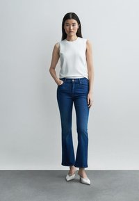 Kvinde stående iført hvid ærmeløs top, mørkeblå vide jeans og hvide hæle mod en ensartet grå baggrund.