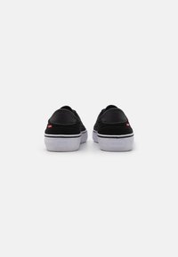 Schwarze Sneaker mit Obermaterial aus Wildleder und Leder, weißer Gummisohle und kontrastierenden schwarzen Nähten, mit einem kleinen roten Etikett an der Ferse.