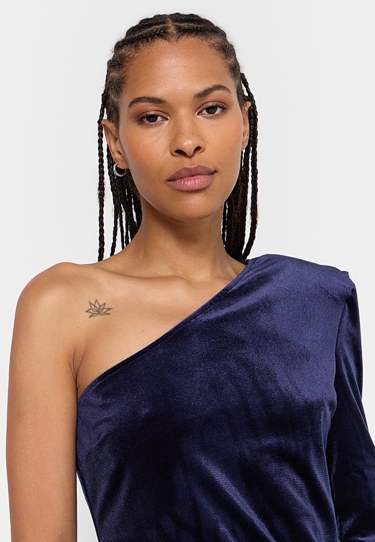 Marineblaues, einseitig trägerloses Kleid aus weichem Samt, das eine glatte Textur und eine taillierte Passform aufweist. Das Modell hat ein florales Tattoo auf ihrer Schulter.