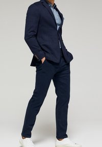 Námořnický blejzr z texturované tkaniny, doplněný světle modrou polo košile a tmavě modrými kalhotami. Dokončeno bílými teniskami.