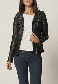 Svart läderbikerjacka med silverdetaljer, dragkedjefickor och en asymmetrisk front. Bärs över en vit skjorta och blå jeans.