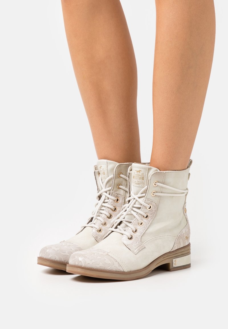 Bottes blanches à lacets avec accents beiges à motifs, portées sur jambes nues, présentant des talons bas et des patches de languette de marque.