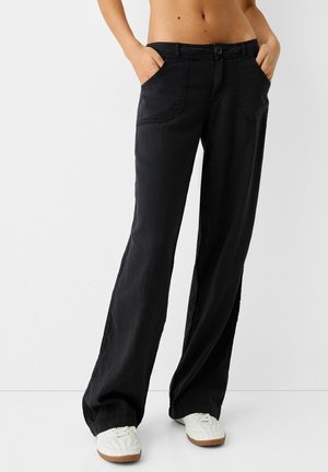 Persona che indossa pantaloni neri larghi con tasche frontali e scarpe da ginnastica bianche, in piedi con le mani nelle tasche contro uno sfondo uniforme.