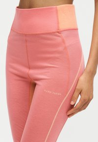 Gros plan d'une personne portant des leggings taille haute roses avec une ceinture pêche contrastante et le logo Kari Traa sur la cuisse, main reposant près de la hanche.