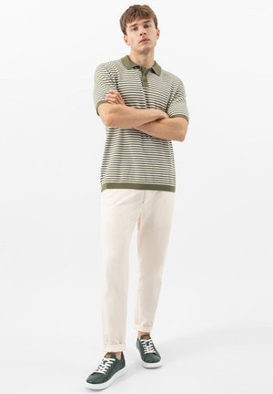 Jeune homme debout, bras croisés, portant un polo rayé vert et blanc, un pantalon crème et des baskets vert foncé avec des lacets blancs.