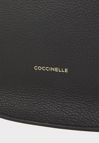Juodos tekstūruotos odos paviršius su subtiliu grūdų raštu, kuriame apačioje dešiniajame kampe įspaustas auksinis "Coccinelle" logotipas.