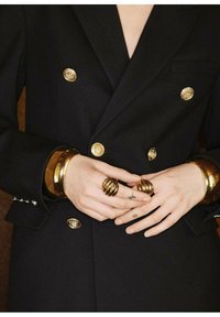 Zwarte blazer met gouden knopen, met een op maat gemaakt ontwerp. Handen versierd met gouden armbanden en spiraalringen, die ingewikkelde texturen onthullen.