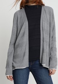 Ljusgrå långärmad cardigan i mjukt tyg med öppen front och subtil ribbad textur. Stylar över en svart topp och blå jeans.
