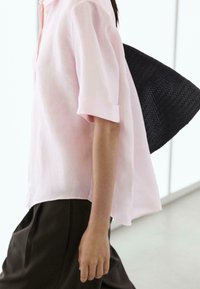 Personne portant une chemise rose clair à manches courtes et un pantalon foncé, portant un grand sac noir tissé sur l'épaule.