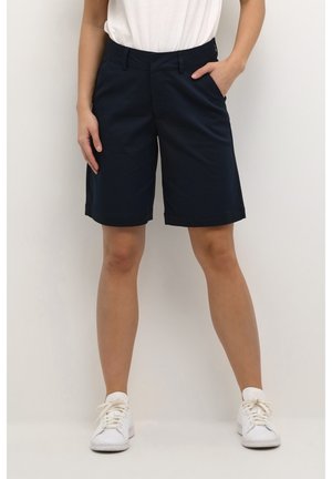 Person iført knælange sorte shorts, en hvid top og hvide sneakers, stående med den ene hånd i lommen mod en ensfarvet baggrund.