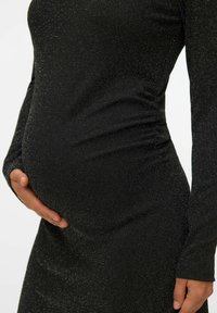 Zwarte, aansluitende jurk met lange mouwen, voorzien van een gestructureerde, glanzende stof en geplooide details rond de taille, die een ronde vorm benadrukken.