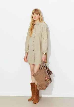 Femme aux longs cheveux blonds portant une robe beige à manches longues boutonnée, des bottes en suède marron et tenant un grand sac cabas marron.