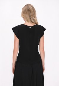 Top nero con maniche a cappotto, texture liscia e una leggera lucentezza. Presenta una cucitura sul retro discreta e un piccolo accento metallico vicino al collo.