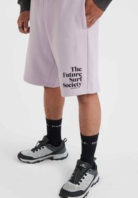Pantalones cortos deportivos de color lavanda claro con texto gráfico negro "The Future Surf Society" en la pierna izquierda, combinados con calcetas negras y zapatos grises.