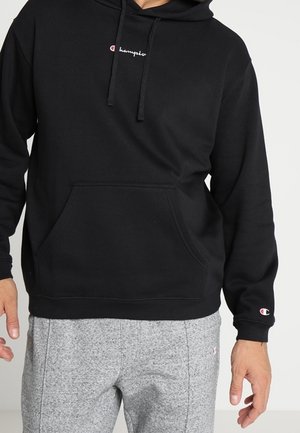 Schwarzer Champion-Hoodie mit Fronttasche und Logo auf Brust und Ärmel, getragen mit hellgrauen Jogginghosen.