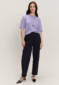 Vrouw die tegen een effen achtergrond staat en een lila korte mouw trui, marineblauwe broek, zwarte hakken en een hartvormige ketting met hanger draagt.