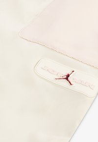 Bela tkanina sročno-belega odtenka s pink panelom, z dvignjenim belim logotipom Jordan in rdečo silhueto Jumpman. Gladka tekstura.
