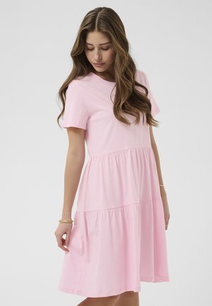 Frau trägt ein rosa, knielanges Kleid mit kurzen Ärmeln und gerafften Stufen, blickt nach unten und hält sanft den Saum des Kleides.