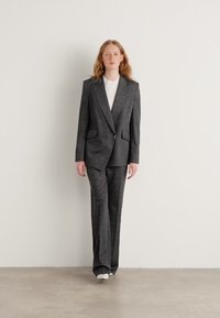 Costume grisé ajusté avec un blazer à boutonnage simple, des revers en pointe, et un pantalon à jambes larges, porté sur une chemise blanche, le tout sur un fond uni.