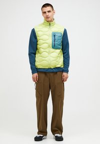 Helles grünes gesteppte Weste mit einer blauen Reißverschlusstasche, getragen über einem blauen Langarm-Oberteil, kombiniert mit braunen Cargo-Hosen und schwarzen Schuhen.