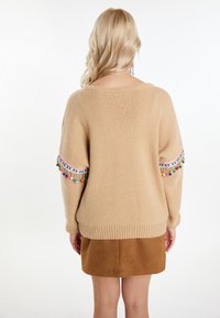 Beige gebreide trui met kleurrijke pompon-accenten op de mouwen. Loose fit, ronde halslijn en geribbelde zoom. Gecombineerd met een bruine rok.