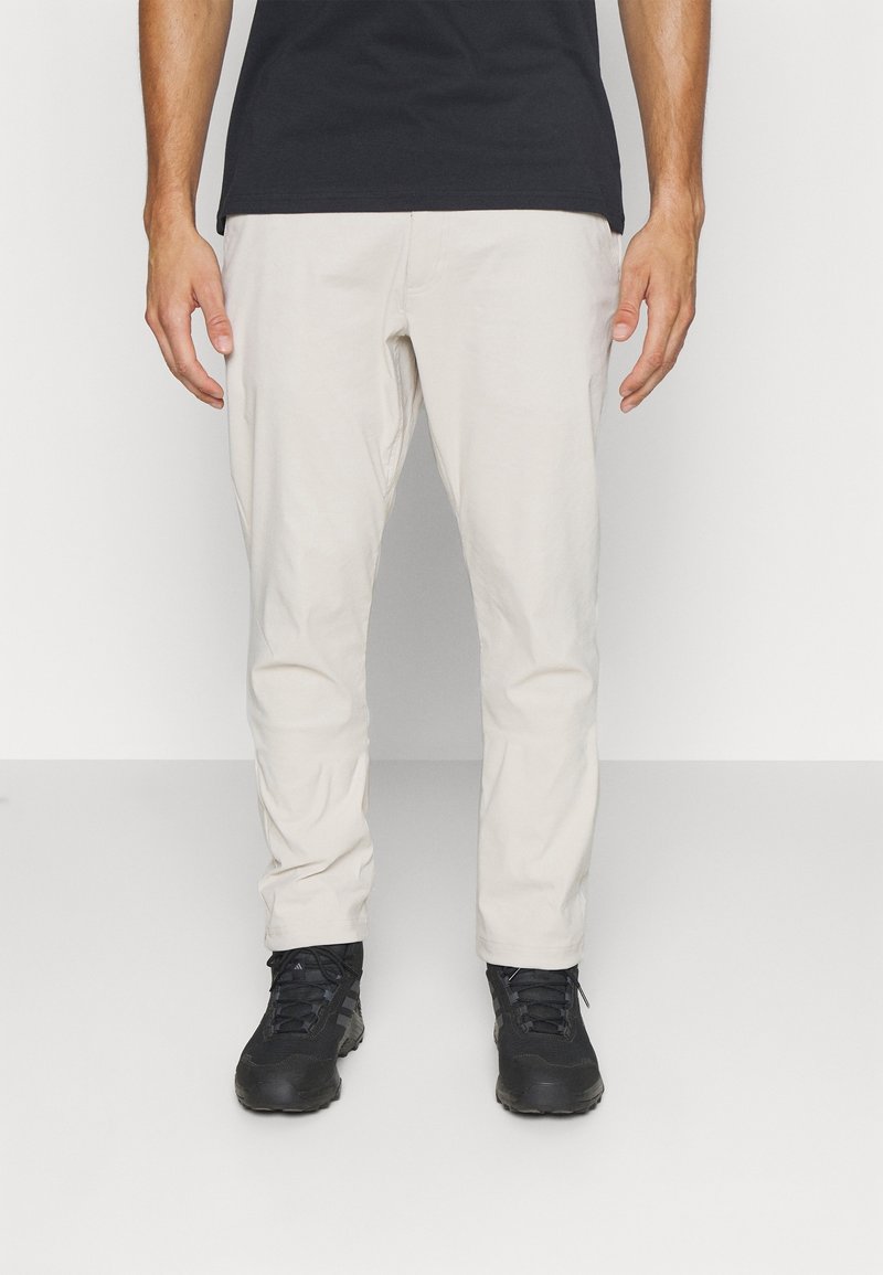 Homme portant un pantalon beige, un t-shirt noir et des baskets noires, debout devant un fond gris clair uni.