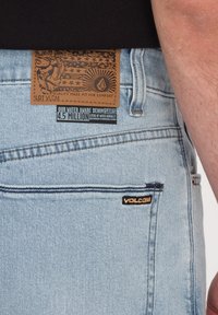 Helle, blaue Jeans aus Denim mit einem strukturierten Lederpatch, das einen Löwen und Sterne zeigt. Enthält ein schwarzes Volcom-Logo auf der Tasche und ein umweltfreundliches Etikett.