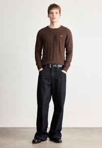 Καφέ πουλόβερ με πλέξη καλωδίων και στρογγυλή λαιμόκοψη, συνδυασμένο με σκούρα μπλε παντελόνια με φαρδιά πόδια και μαύρη ζώνη. Μαύρα παπούτσια ολοκληρώνουν το σύνολο.