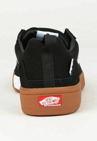 Scarpe da skate nere con tomaia in tela, dettagli bianchi e suola in gomma marrone. Presentano un'etichetta sul tallone con il marchio per facilitarne l'indossamento.